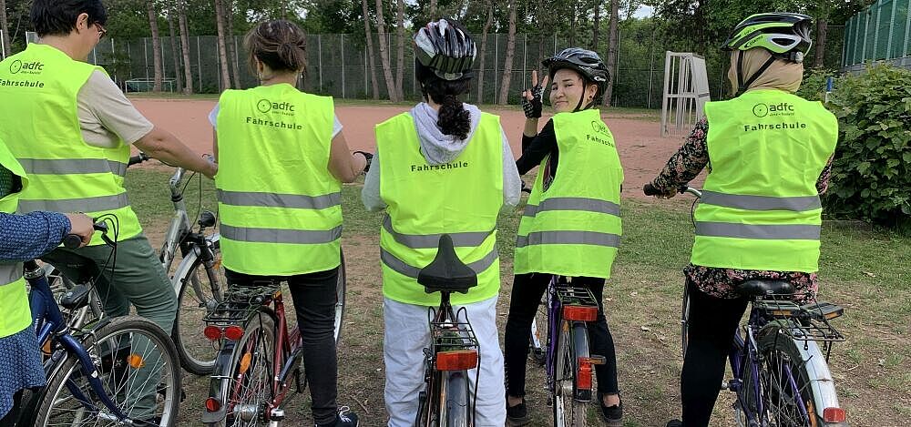 Rückenansicht von stehenden Fahrradfahrerinnen in Sicherheitswesten, eine Frau wendet sich um zeigt mit ihren Fingern das Peace-Zeichen