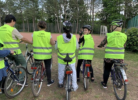 Rückenansicht von stehenden Fahrradfahrerinnen in Sicherheitswesten, eine Frau wendet sich um zeigt mit ihren Fingern das Peace-Zeichen