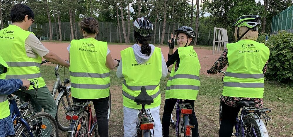 Frauenfahrradschule Rückenansicht von stehenden Fahrradfahrerinnen in Sicherheitswesten, eine Frau wendet sich um zeigt mit ihren Fingern das Peace-Zeichen