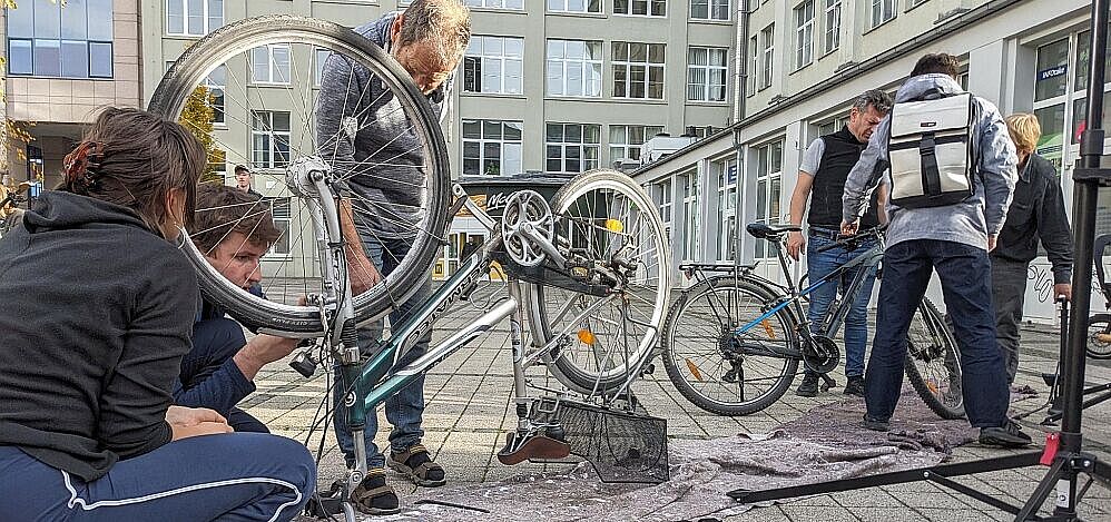 Fahrrad steht mit Reifen nach oben auf einer Decke, während drei Menschen es reparieren