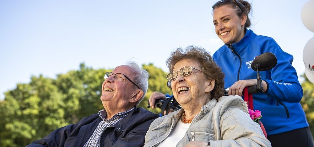Zwei ältere Personen werden von einer jungen Frau in einer Rikscha gefahren. Alle lachen entspannt.