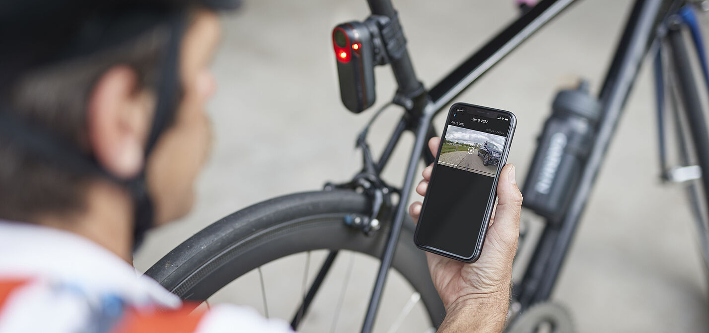Mann kniet mit Smartphone in der Hand vor einen Rennrad mit rot leuchtendem Rücklicht an Sattelstütze. Das Rücklicht Garmin Varia RCT715 mit integrierter Kamera zeichnet permanent auf und speichert Daten aber nur bei einem erkannten Unfall.  